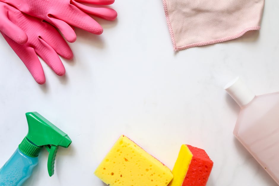 A flat lay of cleaning tools and supplies for surface cleaning and sanitisation, featuring bright pink rubber gloves, a green spray bottle with a blue trigger, a yellow and red sponge, a light pink spray bottle, and a pink cleaning cloth. The items are arranged on a white surface, indicating a setup for domestic or commercial cleaning, with a focus on hygiene and maintenance. This image aligns with Putney Cleaners' expertise in detailed and professional cleaning services in residential flats along Upper Richmond Road, as highlighted in the context of the page 'Cleaning Upper Richmond Road Flats: Local Tips, PUTNEY' available at putney-cleaners.org.uk, ensuring a clear visual representation of cleaning processes.