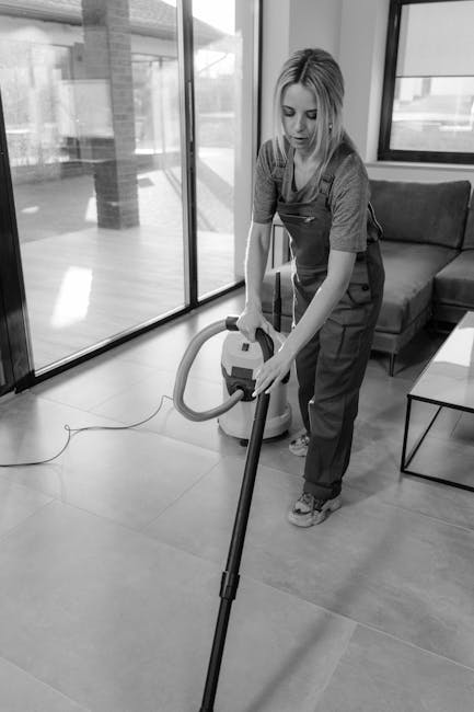 Two professional cleaners from Putney Cleaners are performing domestic surface cleaning in a modern kitchen. One cleaner, a woman, is using a vacuum cleaner on the light-colored wooden floor near a di