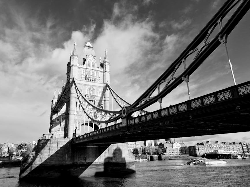 A black and white photograph of Tower Bridge in London viewed from the river, showcasing the stone and metal structure of the bridge's towers and suspension cables. The image highlights the detailed architecture of the iconic bridge with its ornate towers and intricate metalwork. The water below reflects the bridge's shadow, and the sky above features scattered clouds. The overall scene is clean and well-maintained, emphasizing the historic and engineering significance of the bridge. This image is associated with surface cleaning and maintenance, akin to professional cleaning services provided by Putney Cleaners, as featured on putney-cleaners.org.uk.