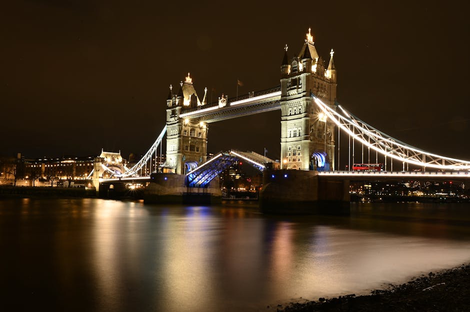 A black and white photograph of Tower Bridge in London viewed from the river, showcasing the stone and metal structure of the bridge's towers and suspension cables. The image highlights the detailed architecture of the iconic bridge with its ornate towers and intricate metalwork. The water below reflects the bridge's shadow, and the sky above features scattered clouds. The overall scene is clean and well-maintained, emphasizing the historic and engineering significance of the bridge. This image is associated with surface cleaning and maintenance, akin to professional cleaning services provided by Putney Cleaners, as featured on putney-cleaners.org.uk.