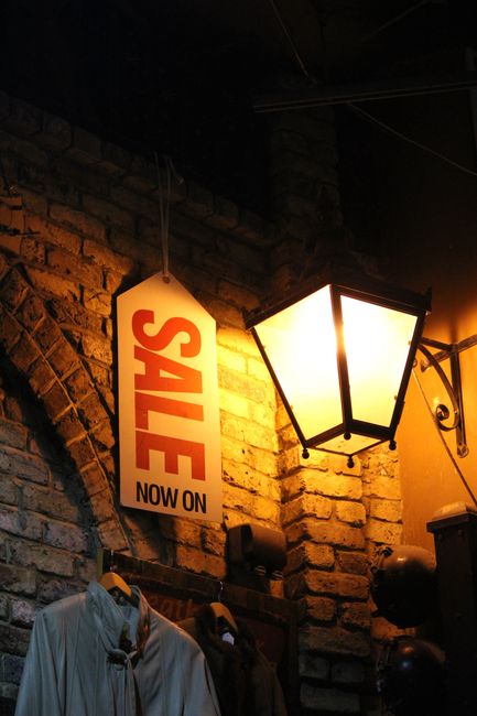 A weathered outdoor advertising box mounted against a red brick wall, featuring a yellow 'On Sale Here' sign with black lettering, and a vertical white 'News Agen' sign attached beside it. The box, po